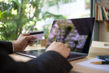 Rear view of modern businessman hands holding credit card typing numbers on computer keyboard while sitting with pen, Calculator on wooden table, soft focus, flare sun light. Online shopping concept.