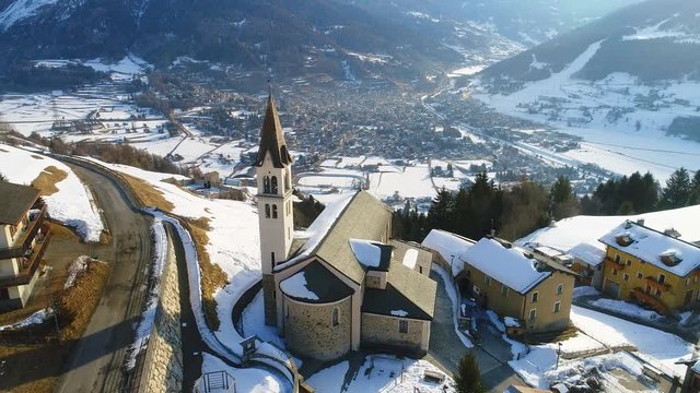 Aerial view of church in mountain village Oga in winter, mountains (Italian Alps) covered with snow, Bormio valley on background, sunrise time - Italy from above, 4k UHD