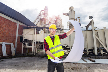 Young engineer or worker wearing yellow safety helmet and vest holding construction plan