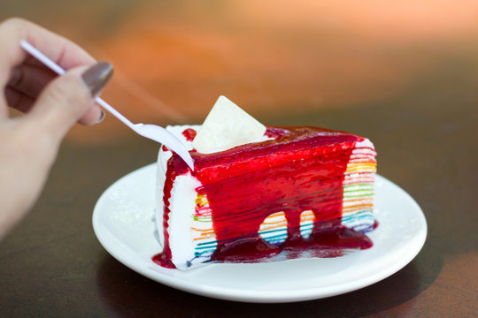 Woman hand hold spoon cutting rainbow crepe cake with Strawberry Sauce
