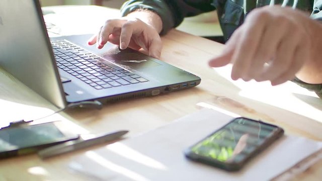 Silhouette of cropped shot of a young man working from home using smart phone and notebook computer, man's hands using smart phone in interior, man at his workplace using technology, flare light