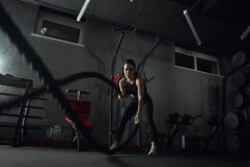 Fototapeta na wymiar Young woman doing strength training using heavy ropes at the gym. Athlete moving the ropes in wave 