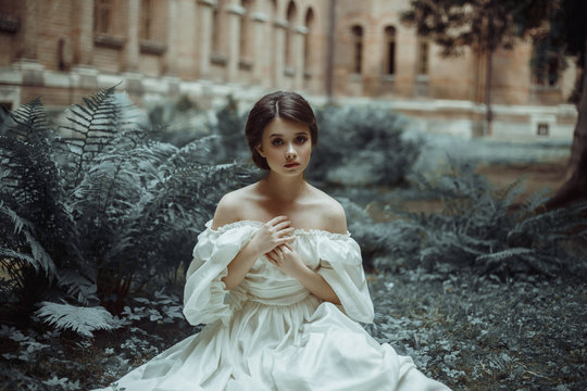 An incredibly beautiful princess sits in the castle garden amid the fern and moss. A beautiful, frightened face. Big sad eyes. Collected hair - a neat hair. On the lady - an old ivory dress. Art photo