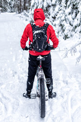 Fototapeta na wymiar A guy in a red jacket is standing with his back on a bicycle in a snowy forest