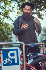 A handsome hipster traveler with a stylish beard and tattoo on his arms dressed in casual clothes and hat, using the phone, standing on the bicycle parking.