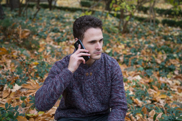 Young handsome man sitting on meadow with foliage and communicating with smartphone. 