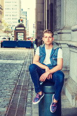 Young American Man with little beard, wearing blue Denim hoody sleeveless vest jacket, white T shirt, jeans, fashionable shoes, sitting on metal pillar on vintage street in New York, relaxing..