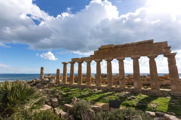 Fototapeta na wymiar SICILY TRAPANI SELINUNTE GREEK TEMPLE ACROPOLIS. Series of columns, at the ancient greek temple over the sea. Archeological site of Selinunte, during a sunny day, with blue sky, clouds and green grass