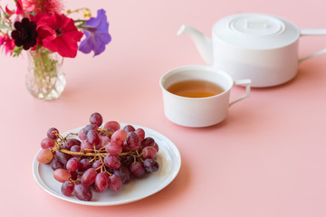 Obraz na płótnie Canvas Red grapes, teacup, teapot and colourful flowers on pink background (selective focus)