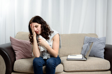 Pretty sad freelancer woman suffering from headache sitting on sofa with laptop. Negative feeling sick, migraine.