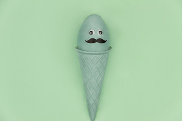 Happy Easter concept. Blue easter egg with funny face and mustache in waffle cone on tint green background