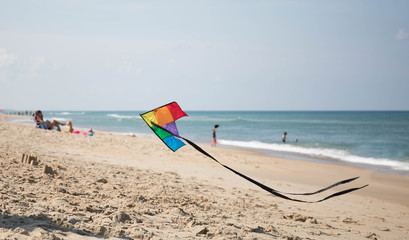 Fototapeta na wymiar A rainbow colored kite flying low over the beach with families and vacationers in the background.