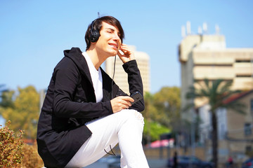 The guy gay is sitting on the bench, using a smartphone to listen to music on headphones. Joyful attractive guy listening to music on headphones while enjoying sitting in the park. Lifestyle concept