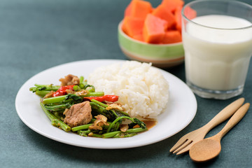 Thai food, stir fried morning glory (Pad Pak Boong) with cooked rice on dish, glass of milk and piece of ripe papaya