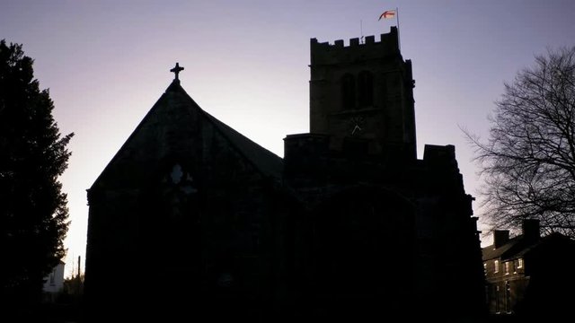 Old sandstone church in shadow illuminated from behind by sunset light. English flag on top of tower. 