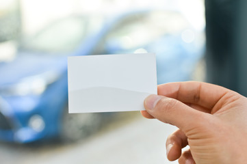 Obraz premium Close up on hand of a businessman holding white business card over blurry car office background. Professional person showing introduction