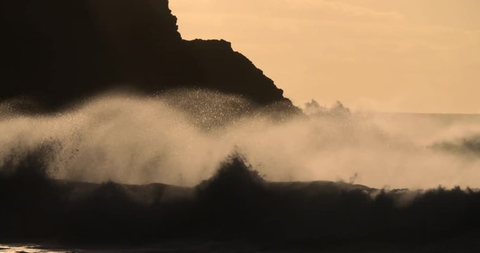 Massive waves break against a rocky shore in golden light in slow motion.