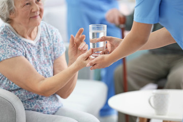 Young caregiver giving glass of water to senior woman indoors