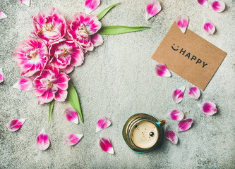 Spring morning concept. Flat-lay of cup of coffee surrounded with pink tulip flowers, petals and sign happy on paper over grey marble background, top view, copy space