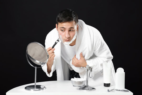 Young man shaving in front of mirror on dark background