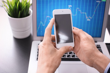 Man using smartphone near laptop with stock chart data on screen. Forex concept