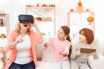 Surprised adult woman in virtual reality glasses sits on couch next to her granddaughter and grandson, who holds tablet.
