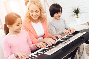 Lovely elderly woman teaches small grandchildren to play synthesizer.
