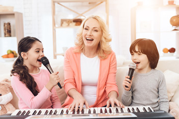 Beautiful elderly woman plays on keyboard with grandchildren who sing into microphone.