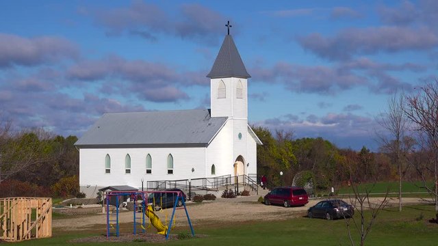 Churchgoers arrive at a pretty white church in the countryside.