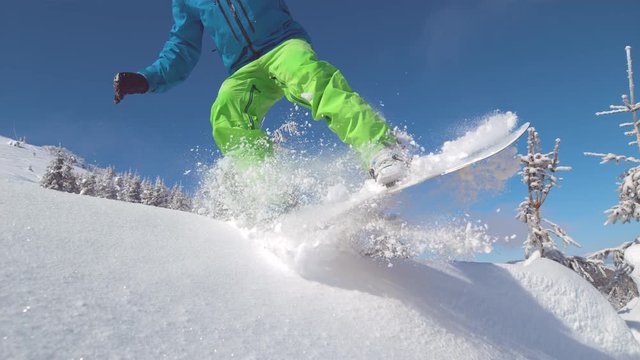 SUPER SLOW MOTION, CLOSE UP: Unrecognizable pro snowboarder jumps into the air while riding downhill on a beautiful winter day. Freerider carving through the sunny backcountry jumps in powder snow.