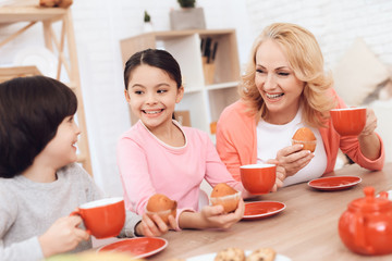 Elderly woman with cheerful grandson and granddaughter eating cookies and drinking tea in red mugs at kitchen.