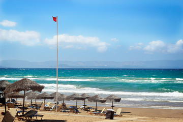 Greece. Red warning flag on beach. Straw beach umbrellas on stormy day against the sea.