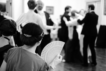 Two women in vintage dresses with fan in hands sitting at ball and looking at couples dancing on...