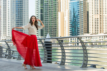 Attractive, Slim, Beautiful And Brunette Girl Wearing White Shirt And Maxi Long Red Silk Fabric Skirt Fly And Wave In The Wind Walking Alongside Dubai Marina Middle East Fashion Concept