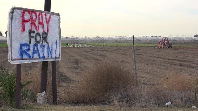 A tractor plows a very dry field during a time of drought in California while a sign urges people to pray for rain.