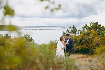 Beautiful wedding photosession. Handsome groom in blue formal suit and bow tie with boutonniere and his elegant bride in white dress and veil with a beautiful hairdress on a walk in field near river