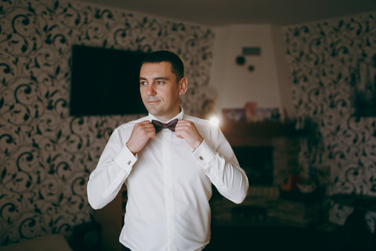 Morning preparation of the newlyweds for the wedding ceremony. The groom in a white shirt with a bracelet on his hand dresses a patterned bow tie on his collar and smiles. Wedding wear, accessories