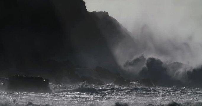 Huge ocean waves roll and crash into a rocky shore in slow motion.