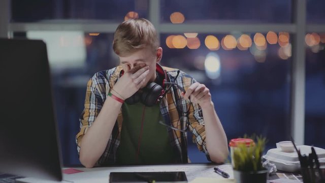 A young designer or a novice intern works in an empty office at a later time, feels tired and rubs his eyes. Workaholic man stayed up late at work, tired of the computer monitor.