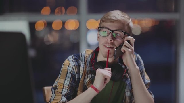 Young man speaks on the phone at the computer in front of a window. Creative man is working in late hours at office and talking on phone. Freelancer works late at night, distracted by a phone call