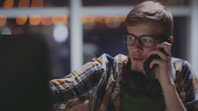 Young man speaks on the phone at the computer in front of a window. Creative man is working in late hours at office and talking on phone. Freelancer works late at night, distracted by a phone call