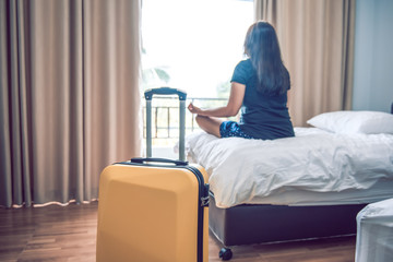 Yellow travel suitcase in the bedroom with woman sitting meditation on the bed and window curtain...