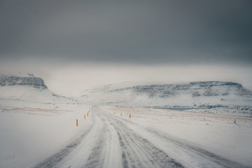 Winter landscape of Iceland's road during snowstorm with the snow-capped mountain as a background and frozen road as a foreground.
