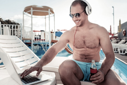 No Time For Worries. Relaxed Young Man Looking At A Screen Of His Laptop With A Cheerful Smile On His Face While Drinking A Bottle Of Sparkling Water And Sunbathing Alone.