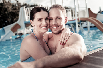 Fototapeta na wymiar Love is in the air. Beautiful young woman hugging her boyfriend tightly while both standing in a swimming pool and grinning broadly into the camera.