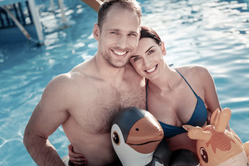 Fototapeta premium Positive vibes. Top view on a cheerful millennial woman and man looking into the camera with smiles on their faces while both wearing colorful rubber rings in a pool.