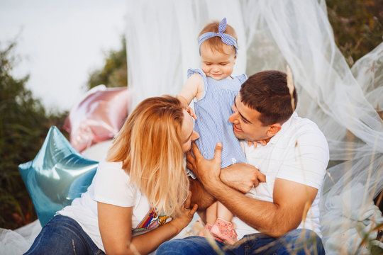 Happy mom and dad hold little daughter on their arms sitting under white tent