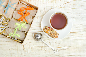 Obraz na płótnie Canvas Mug of tea, muesli bars and tea strainer. Box with bars. White wooden background