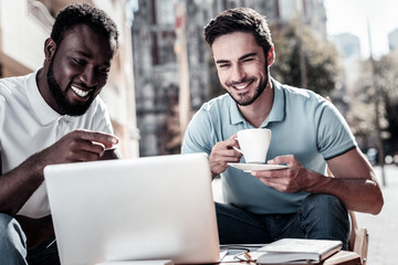 What do you think. Happy young men smiling cheerfully while sitting next to each other and looking at a laptop during a work session outdoors.