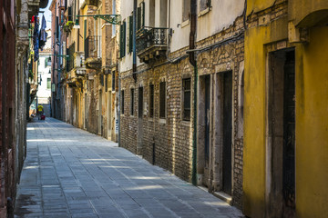 Narrow street in Venice, Italy, a tourist with red suitcase in background. Architectural details of old houses and historic buildings.